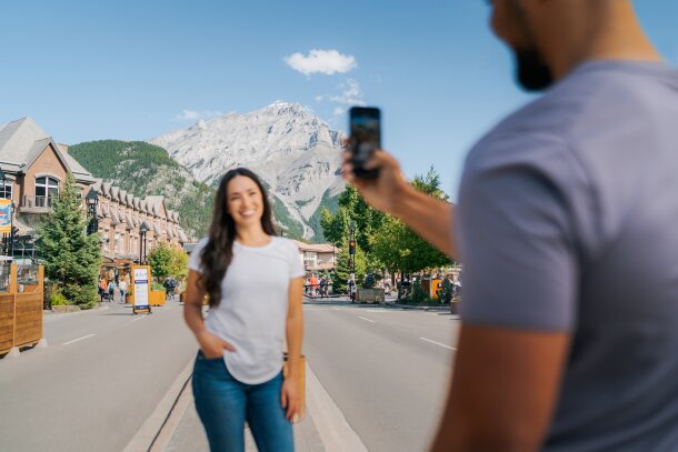 A couple takes a photo on the Banff Avenue Pedestrian Zone with Cascade Mountain in the background