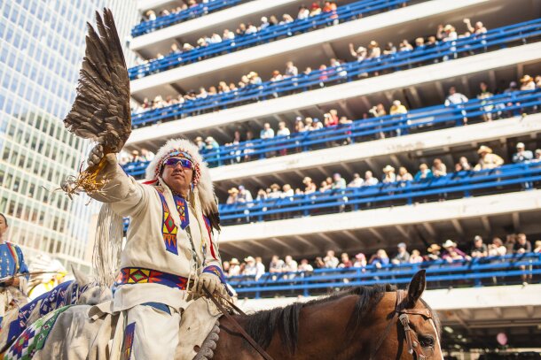 First Nations man on horse riding in front of parking garage lined with spectators